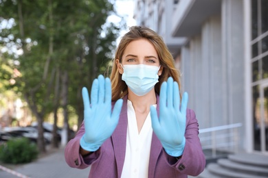 Woman in protective face mask showing stop gesture outdoors. Prevent spreading of coronavirus