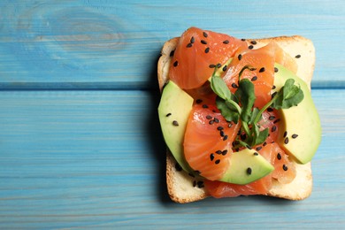 Tasty toast with butter, salmon, avocado, sesame seeds and microgreens on light blue wooden table, top view. Space for text
