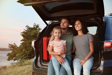 Happy young family sitting in car trunk on riverside
