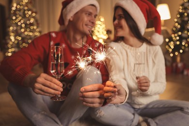 Couple in Santa hats holding sparkles and champagne glasses, focus on fireworks. Christmas celebration