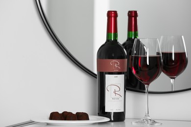 Bottle and glass of red wine with chocolate candies on table near mirror
