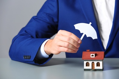 Man covering house model with umbrella cutout at table, closeup. Home insurance