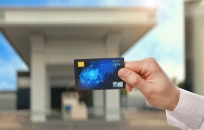 Man holding credit card against blurred gas station, closeup. Cashless payment