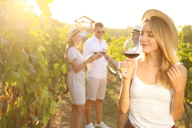 Beautiful young woman with glass of wine and her friends at vineyard on sunny day