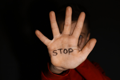 Photo of Abused little boy showing palm with word STOP against black background, focus on hand. Domestic violence concept