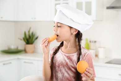 Cute little girl wearing chef hat eating cookies in kitchen