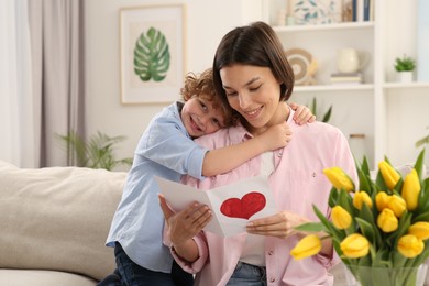 Photo of Little son congratulating his mom with Mother`s day at home. Woman holding handmade greeting card