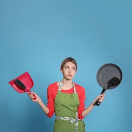 Young housewife with dustpan, brush, frying pan and spatula on light blue background