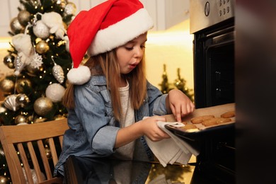 Little child in Santa hat taking baking sheet with Christmas cookies out of oven indoors