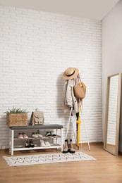 Stylish hallway interior with coat rack, shoe storage bench and mirror near white brick wall