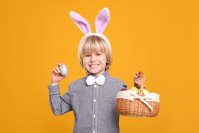 Photo of Happy boy in bunny ears headband holding wicker basket with painted Easter eggs on orange background