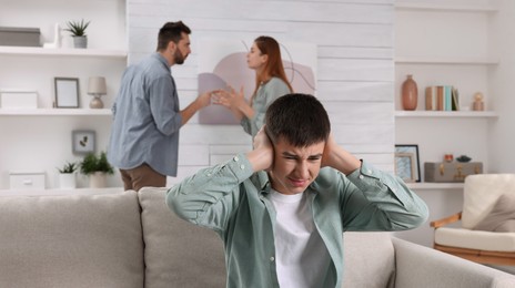 Unhappy teenage boy covering ears while his parents arguing on background. Problems at home