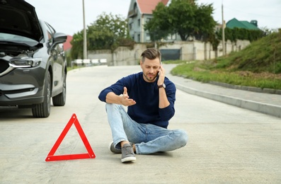 Man talking on phone near warning triangle and broken car on road