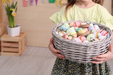 Little girl holding wicker basket full of Easter eggs indoors, closeup. Space for text