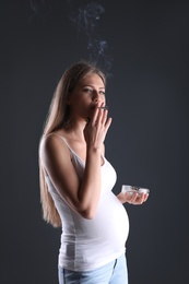 Young pregnant woman smoking cigarette on dark background. Harm to unborn baby
