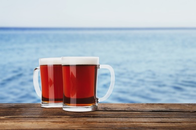 Glass mugs of delicious kvass on wooden table near sea. Space for text