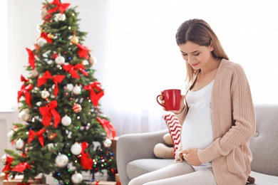 Happy pregnant woman with cup of tea sitting on sofa in room decorated for Christmas