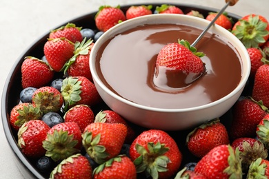 Fondue fork with strawberry in bowl of melted chocolate surrounded by different berries on light table, closeup