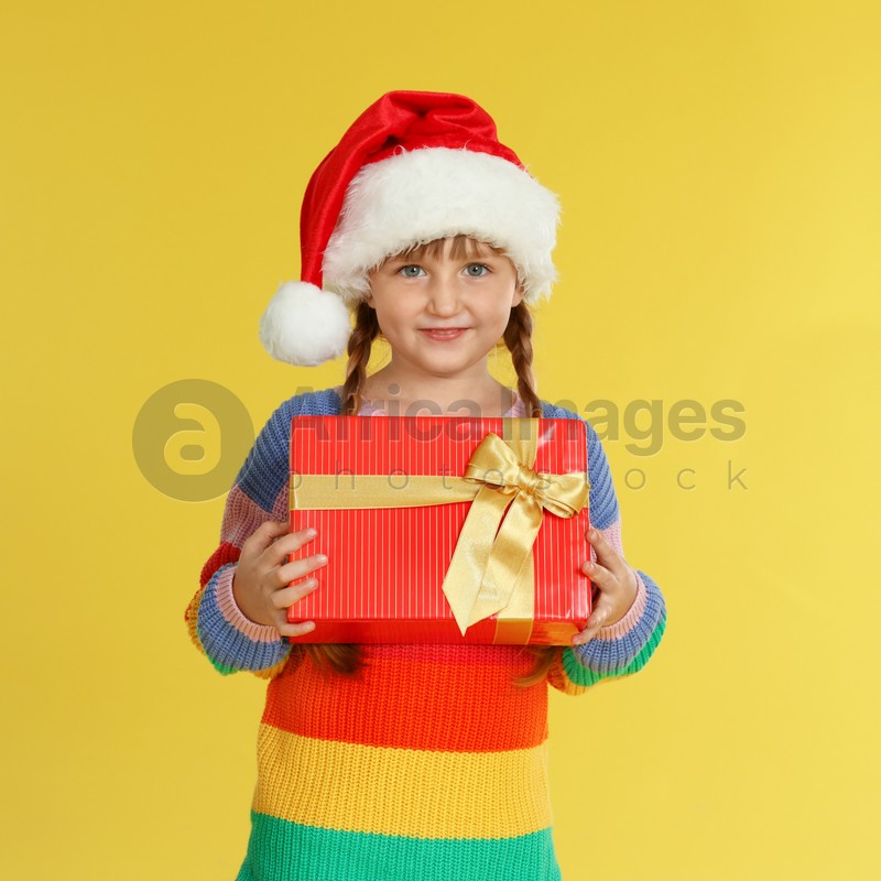 Cute little girl in Santa hat with Christmas gift on yellow background