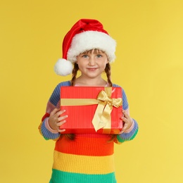 Cute little girl in Santa hat with Christmas gift on yellow background