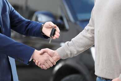 Photo of Salesman giving key to customer while shaking hands in modern auto dealership, closeup. Buying new car