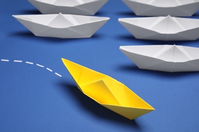 Photo of Yellow paper boat floating away from others on blue background, closeup. Uniqueness concept
