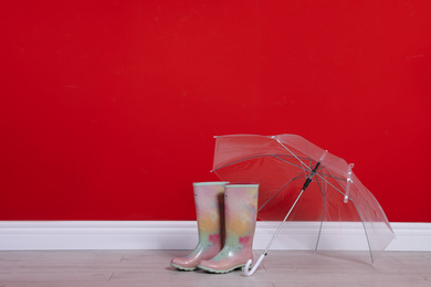 Beautiful transparent umbrella and rubber boots near red wall. Space for text