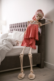 Human skeleton in silk pajamas and towel sitting on bed indoors