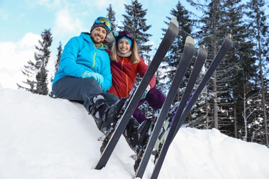 Happy couple with ski equipment sitting on snowdrift outdoors. Winter vacation