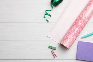 Roll of festive wrapping paper, ribbon, card, pen and clothespins on white wooden table, flat lay. Space for text