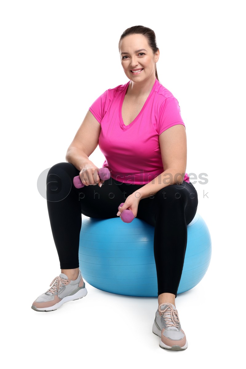 Happy overweight woman with dumbbells sitting on fitness ball against white background