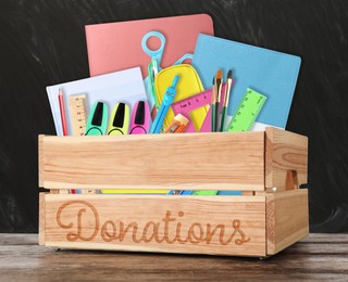 Donation box with different school stationery on wooden table near blackboard