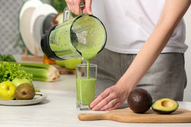 Woman pouring tasty green smoothie into glass at white wooden table, closeup