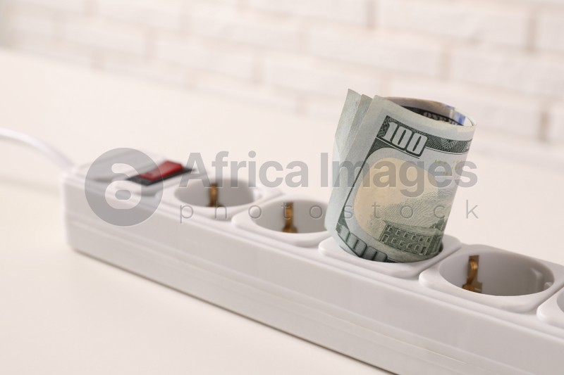 Extension board with rolled dollar banknotes on white table near brick wall, closeup