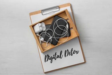 Different gadgets in crate and clipboard with words DIGITAL DETOX on white wooden background, top view