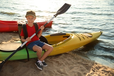 Happy boy with paddle sitting on kayak near river. Summer camp activity