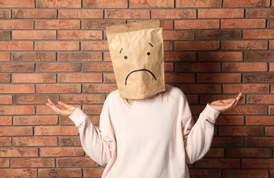 Woman wearing paper bag with drawn sad face near brick wall