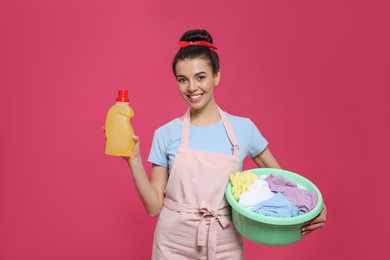 Housewife holding bottle of cleaning product and basin with clothes on pink background