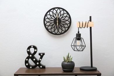 Photo of Beautiful plant in pot, lamp and decor on wooden table near light wall with clock indoors. Interior accessories