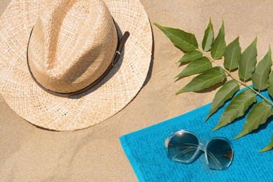 Photo of Soft blue beach towel, sunglasses, straw hat and green leaves on sand, flat lay