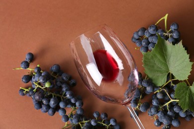 Overturned glass with red wine and grapes on brown background, flat lay
