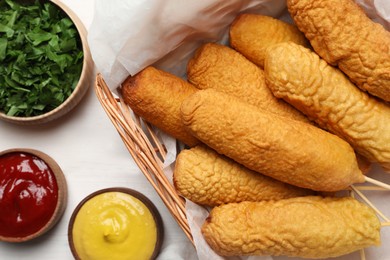 Delicious deep fried corn dogs in basket and sauces on white wooden table, flat lay