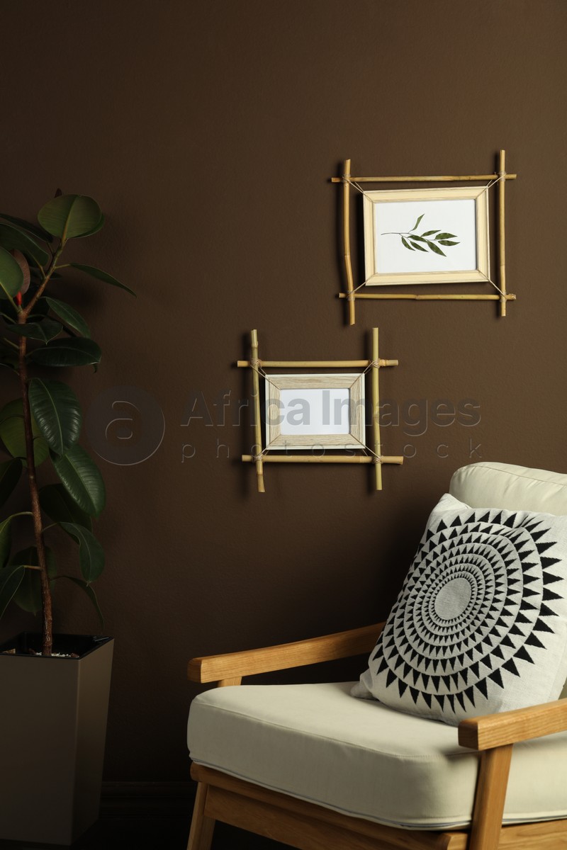 Beautiful pictures in bamboo frames on brown wall and stylish armchair indoors. Interior design