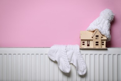 Modern radiator with knitted hat, socks and wooden house near pink wall indoors, space for text. Winter heating efficiency