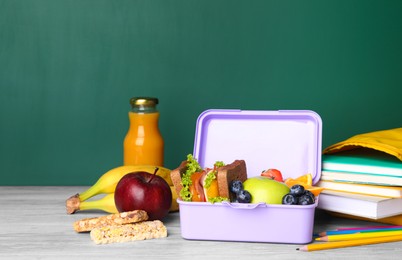 Lunch box with healthy food and different stationery on light wooden table near green chalkboard, space for text