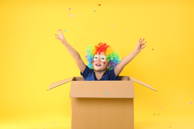 Little boy in clown wig sitting inside of cardboard box under confetti shower on yellow background. April fool's day