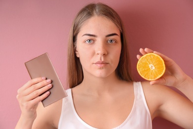 Young woman with acne problem holding orange and chocolate bar on color background. Skin allergy