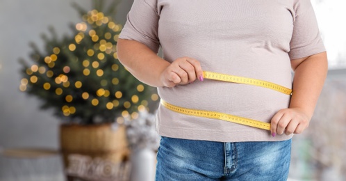 Overweight woman measuring her waist in room decorated for Christmas after holidays, closeup