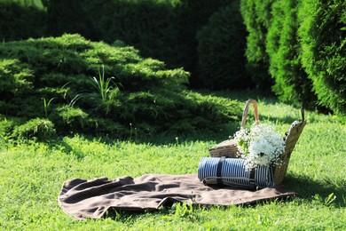 Picnic blanket with beautiful flowers, rolled mat and basket in garden on sunny day