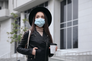 Young woman in medical face mask with cup of coffee walking outdoors. Personal protection during COVID-19 pandemic